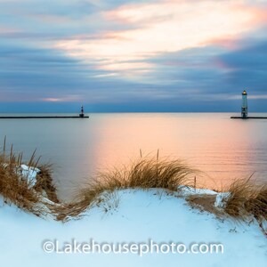 Puede incluir: Dos faros se alzan sobre muelles que se adentran en una masa de agua. El agua está tranquila y refleja los tonos rosados y anaranjados del cielo al atardecer. En primer plano, una duna cubierta de nieve con hierbas marrones.