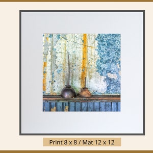 May include: Two vintage oil cans with long spouts sit on a weathered blue and yellow wood shelf. The cans are rusty and have a worn patina. The background is a peeling blue and yellow wall.