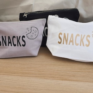 May include: Two canvas zippered pouches with the word "SNACKS" printed on them. One pouch is light grey with black text and a cookie graphic. The other pouch is white with gold text and a cookie graphic.