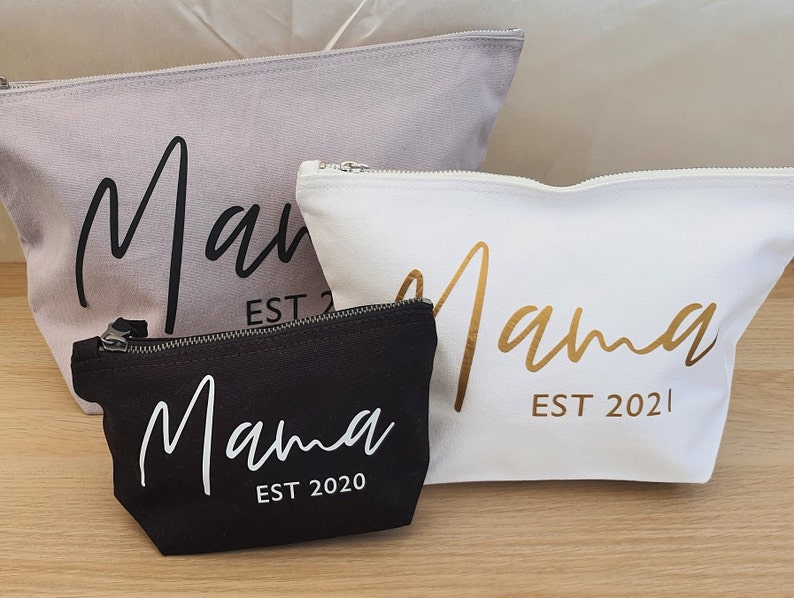 Puede incluir: Tres estuches con cremallera con la palabra "Mama" en diferentes fuentes y colores. Los estuches son grises, blancos y negros. El texto en el estuche gris es negro, el texto en el estuche blanco es dorado y el texto en el estuche negro es blanco. Cada estuche tiene el a&ntilde;o "EST 2020" o "EST 2021" impreso debajo de la palabra "Mama".
