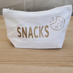 May include: White canvas zippered pouch with a gold outline of a cookie and the word "SNACKS" in gold lettering.