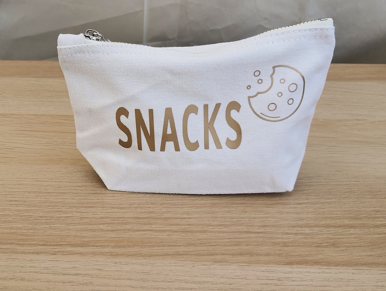 May include: White canvas zippered pouch with a gold outline of a cookie and the word "SNACKS" in gold lettering.