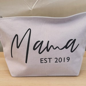 Puede incluir: Una bolsa de lona gris claro con cremallera con la palabra "Mama" escrita en cursiva negra y el texto "EST 2019" debajo.