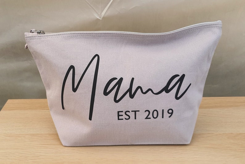 Puede incluir: Una bolsa de lona gris claro con cremallera con la palabra "Mama" escrita en cursiva negra y el texto "EST 2019" debajo.
