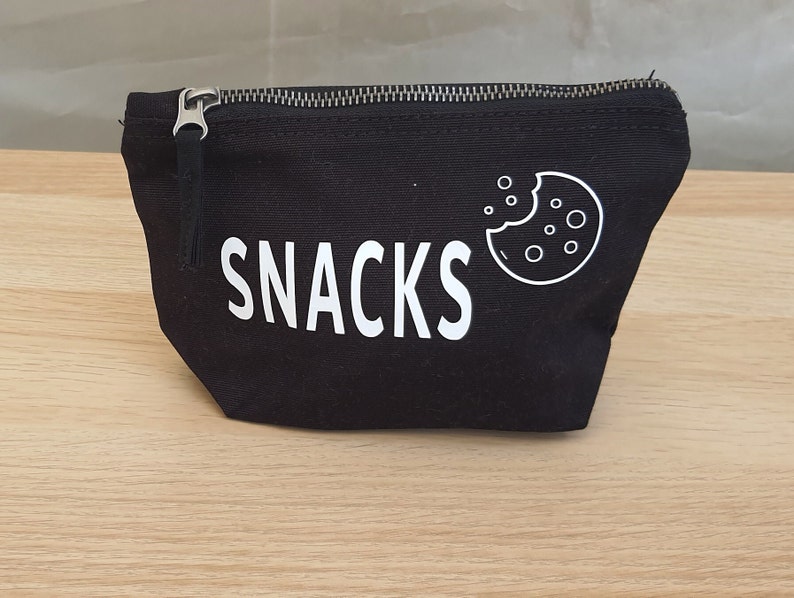 May include: Black canvas zippered pouch with the word "SNACKS" printed in white and a white outline of a cookie with a bite taken out of it.