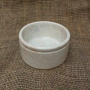 May include: A white ceramic bowl with a crackled glaze. The bowl is round and has a slightly flared rim. It is sitting on a brown burlap surface.