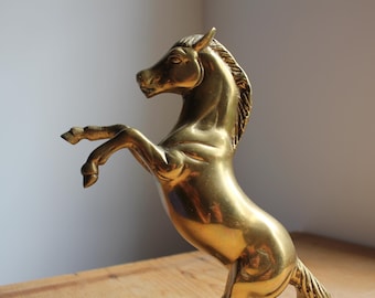 May include: A golden brass horse statue rearing up on its hind legs. The sculpture has a polished finish, catching the light and highlighting the details of the horse's mane and muscular form. A decorative equestrian piece.