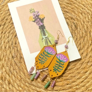 May include: A pair of dangle earrings with a floral design. The earrings are made of leather and feature a purple flower with green leaves. The earrings are accented with purple and green beads.