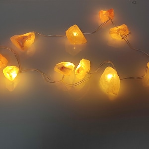 Könnte beinhalten: Eine Lichterkette mit beleuchteten, bernsteinfarbenen Himalaya-Salzsteinen. Die Lichter sind auf einem dünnen, silbernen Draht aufgereiht und strahlen ein warmes, einladendes Leuchten aus. Die unregelmäßig geformten Salzsteine variieren in der Größe.