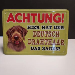 Könnte beinhalten: Ein gelbes Metallschild mit dem Text "ACHTUNG! HIER HAT DER DEUTSCH DRAHTHAAR DAS SAGEN!" in schwarzer und roter Schrift. Ein brauner und weißer Deutsch Drahthaar Hund ist in der oberen linken Ecke des Schildes abgebildet.