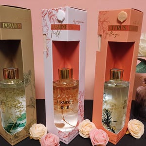 Könnte beinhalten: Drei Glasdiffuserflaschen mit dekorativen Schachteln. Die Schachteln sind mit "Power", "Jasmine Field" und "Citrus Collage" beschriftet. Die Flaschen sind mit einer klaren Flüssigkeit gefüllt und haben ein florales Design.