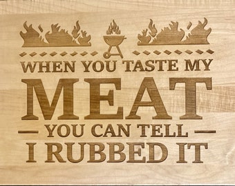 Puede incluir: Una tabla de cortar de madera con un diseño grabado con láser. El diseño presenta llamas, una parrilla y el texto "When you taste my meat, you can tell I rubbed it."