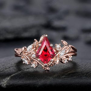 May include: Rose gold ring featuring a kite-shaped, vibrant red gemstone as the centerpiece. The ring is adorned with delicate leaf and vine detailing, and accented with small, clear gemstones. The ring is set on a dark, textured surface.