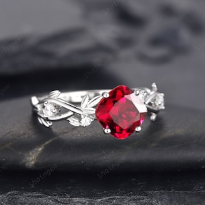 以下が含まれることがあります： 赤い正方形の大きな宝石が、小さな白い宝石で囲まれた銀の指輪。指輪のバンドは、繊細な葉のデザインで飾られています。