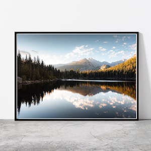 May include: A scenic photograph of a mountain lake with a reflection of the surrounding trees and mountains in the water. The sky is blue with white clouds and the water is calm and still.