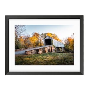 May include: Framed print of a covered bridge in a rural setting. The bridge is white with a weathered appearance, set against a backdrop of vibrant autumn foliage in shades of yellow and orange. The bridge is supported by stone pillars.