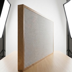 May include: A rectangular, light-colored wooden frame with a gray mesh screen. The frame is made of light-colored wood and sits on a wooden surface. The background is a white studio setup with lighting equipment.