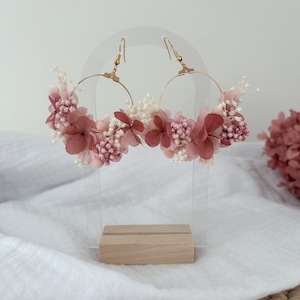 Peut inclure: Une paire de boucles d'oreilles créoles ornées de fleurs séchées roses et blanches disposées autour des cercles. Les boucles d'oreilles sont présentées sur un support en acrylique transparent avec une base en bois.