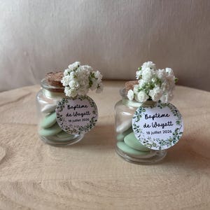 May include: Two small glass jars filled with green and white sweets, topped with corks and white flowers. Each jar has a round label with the text "Baptême de Wayatt" and "18 juillet 2026" in French, surrounded by a green leaf design.