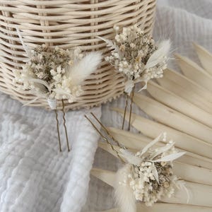 May include: Three floral hair pins with dried white flowers and feathers. The pins are arranged on a white cloth with a woven basket in the background. The hair accessories are ideal for weddings or special occasions.