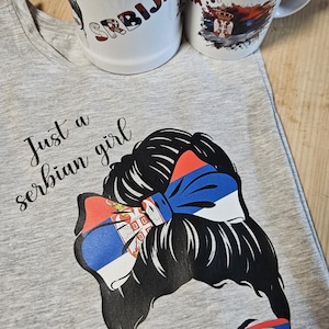Op de afbeelding: Een grijs t-shirt met de tekst "Just a Serbian girl" en een afbeelding van een vrouw met een Servische vlagstrik en bril. Twee witte keramische mokken met Servische vlagontwerpen en het woord "SRBIJA" zijn ook aanwezig.