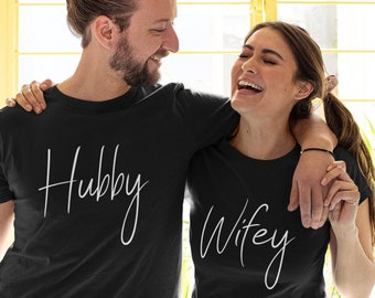 Może przedstawiać: Para w dopasowanych czarnych koszulkach z napisami "Hubby" i "Wifey" w białym kolorze. Uśmiechają się i patrzą na siebie.