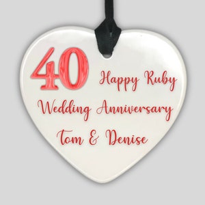 Puede incluir: Adorno de cerámica blanco en forma de corazón con una cinta roja. El adorno tiene el número 40 en rojo, el texto "Happy Ruby Wedding Anniversary Tom & Denise" en rojo.