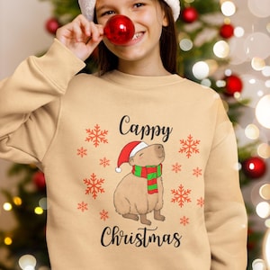 Könnte beinhalten: Beiges Sweatshirt mit dem Text "Cappy Christmas" und einer Grafik eines Capybaras mit Weihnachtsmütze und Schal. Rote Schneeflocken sind um das Design herum verstreut. Eine Person trägt einen Weihnachtsmütze und hält eine rote Kugel.