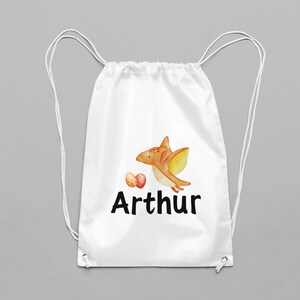 Könnte beinhalten: Weiße Kordeltasche mit einer Cartoon-Illustration eines braunen und gelben Pterodactylus mit zwei roten und orangefarbenen Eiern. Der Text "Arthur" ist unter der Illustration gedruckt.