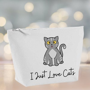 Puede incluir: Bolsa de lona blanca con cremallera con un gato gris de dibujos animados con ojos amarillos y lengua rosa. El texto "I Just Love Cats" está impreso en negro debajo del gato.
