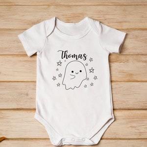 May include: White baby onesie with a cute ghost illustration and the name "Thomas" printed on it. The onesie is surrounded by fall leaves and apples on a wooden background.