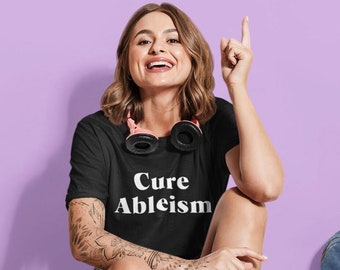 Può includere: Una maglietta nera con le parole "Cure Ableism" in bianco, indossata da una persona seduta su uno skateboard giallo. La persona sorride e indica verso l'alto. Le cuffie sono intorno al collo.