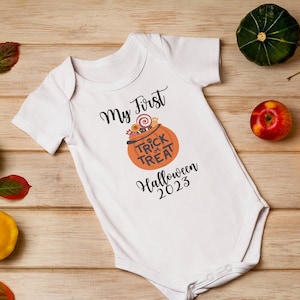 Könnte beinhalten: Ein weißer Baby-Strampler mit dem Text "My First Halloween 2023" und einer Grafik einer Kürbis mit dem Text "Trick or Treat". Der Strampler ist umgeben von Herbstdekorationen, darunter Kürbisse, Äpfel und Blätter.