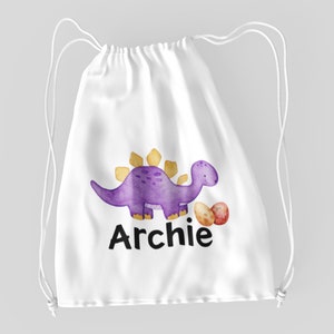 Könnte beinhalten: Weiße Kordeltasche mit einem lila Dinosaurier und goldenen Eiern. Der Dinosaurier hält ein Ei im Maul. Die Tasche ist mit dem Namen "Archie" bedruckt.