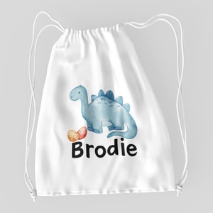 Könnte beinhalten: Weiße Kordeltasche mit einem blauen Dinosaurier und zwei Eiern. Die Tasche ist mit dem Namen "Brodie" bedruckt.