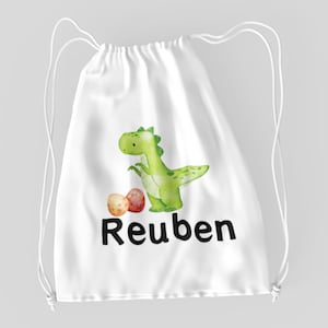 Könnte beinhalten: Weiße Kordeltasche mit einem grünen Dinosaurier und drei Eiern. Die Tasche ist mit dem Namen "Reuben" bedruckt.