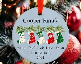 May include: A blue and white Christmas ornament with the text "Cooper Family" and five stockings hanging from a garland. The stockings are labeled "Mum", "Dad", "Jude", "Luca", and "Freya". The ornament also includes the text "Christmas 2024".