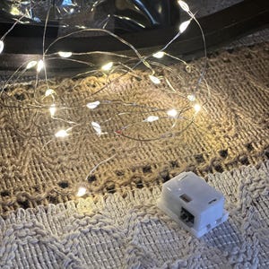 May include: A close-up of a string of small, warm white LED lights on a thin, flexible silver wire. A small, white battery pack is visible. The lights are resting on a textured, off-white and tan fabric.