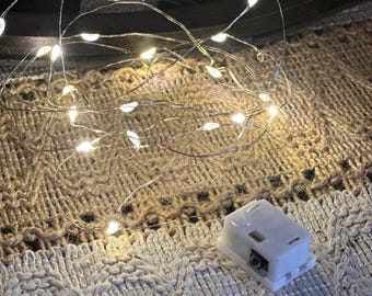 May include: A close-up of a string of small, warm white LED lights. The lights are on a thin, flexible silver wire. A small, white battery pack is visible. The lights are resting on a textured, off-white and tan fabric.