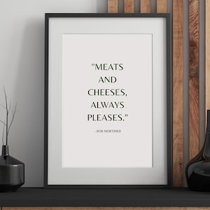 May include: A framed print featuring the quote "MEATS AND CHEESES, ALWAYS PLEASES." by Bob Mortimer. The text is in a serif font on a white background, framed in black. The print is displayed on a black shelf with decorative vases.