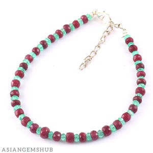 Könnte beinhalten: Ein Armband mit abwechselnd Rubinen und Smaragden. Das Armband hat einen silberfarbenen Verschluss und eine Verlängerungskette. Die Edelsteine sind tiefrot und hellgrün. Das Armband wird vor einem weißen Hintergrund gezeigt.