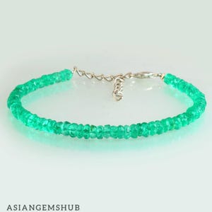 Puede incluir: Una pulsera de piedras preciosas verde vibrante con un cierre plateado. La pulsera está compuesta por pequeñas cuentas redondas de color esmeralda ensartadas. Las cuentas son de un color verde brillante y translúcido. La pulsera se exhibe sobre una superficie blanca.