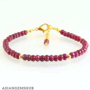 May include: A ruby gemstone bracelet with a gold clasp and accents. The bracelet features small, faceted, deep red ruby beads strung together. The clasp and connecting elements are gold-colored.