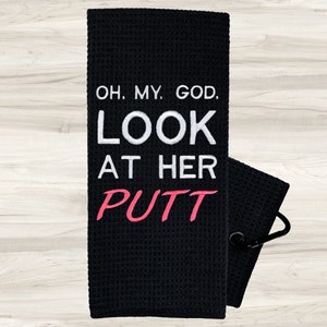 May include: Black golf towel with white text that reads "Oh. My. God. Look at her putt" with the word "putt" in pink.