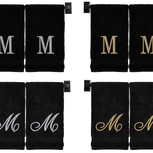May include: Two sets of black hand towels with embroidered letter "M" in different styles. The first set has silver "M"s, the second set has gold "M"s, the third set has silver cursive "M"s, and the fourth set has gold cursive "M"s.