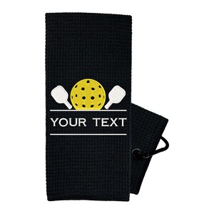 Pode incluir: Toalha de tecido waffle preto com um design bordado branco de duas raquetes de pickleball e uma bola de pickleball amarela. O texto "YOUR TEXT" est&aacute; bordado abaixo do design.