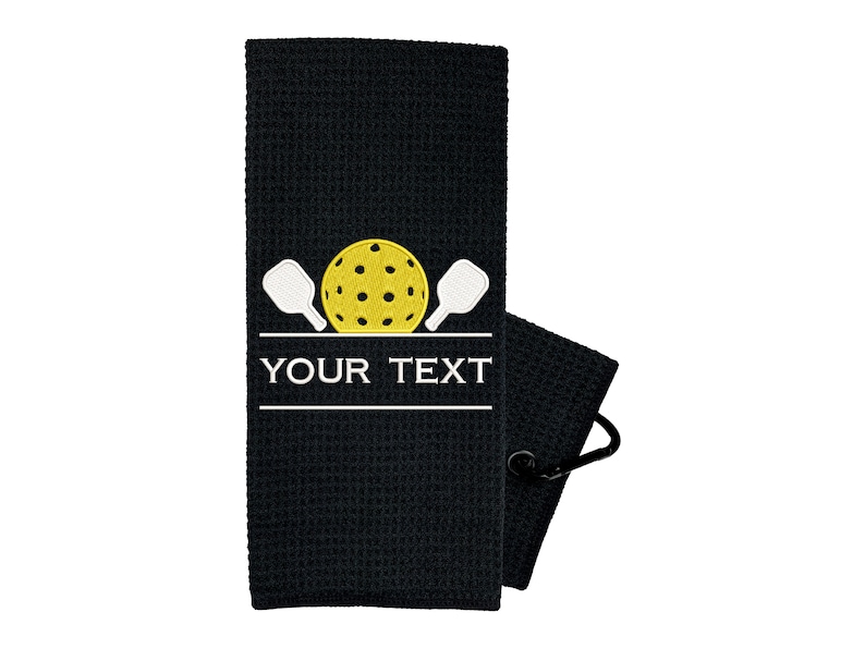 Pode incluir: Toalha de tecido waffle preto com um design bordado branco de duas raquetes de pickleball e uma bola de pickleball amarela. O texto "YOUR TEXT" est&aacute; bordado abaixo do design.