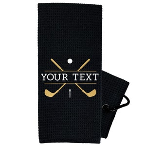 May include: Black golf towel with a white golf club and ball design. The towel has a loop for hanging and the text "YOUR TEXT" in white.