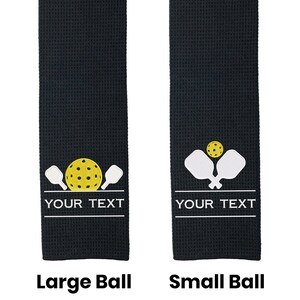 Pode incluir: Duas toalhas de tecido waffle preto com um design branco de duas raquetes de pickleball e uma bola de pickleball amarela. A toalha &agrave; esquerda tem uma bola maior e a toalha &agrave; direita tem uma bola menor. Ambas as toalhas t&ecirc;m o texto "YOUR TEXT" impresso abaixo do design.