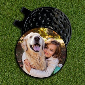 May include: A black golf ball marker with a circular photo of a girl hugging a golden retriever. The marker has a dimpled surface and a handle. The photo shows the girl and dog outdoors.